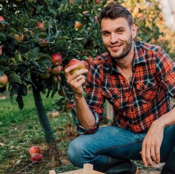 Elma bahçesinde bir çiftçi, sağlıklı ve kırmızı bir elmayı gösteriyor. Bor ve Molibden çözümleri ile giderilmiş mikro besin eksiklikleri sayesinde elde edilmiş kaliteli meyve.